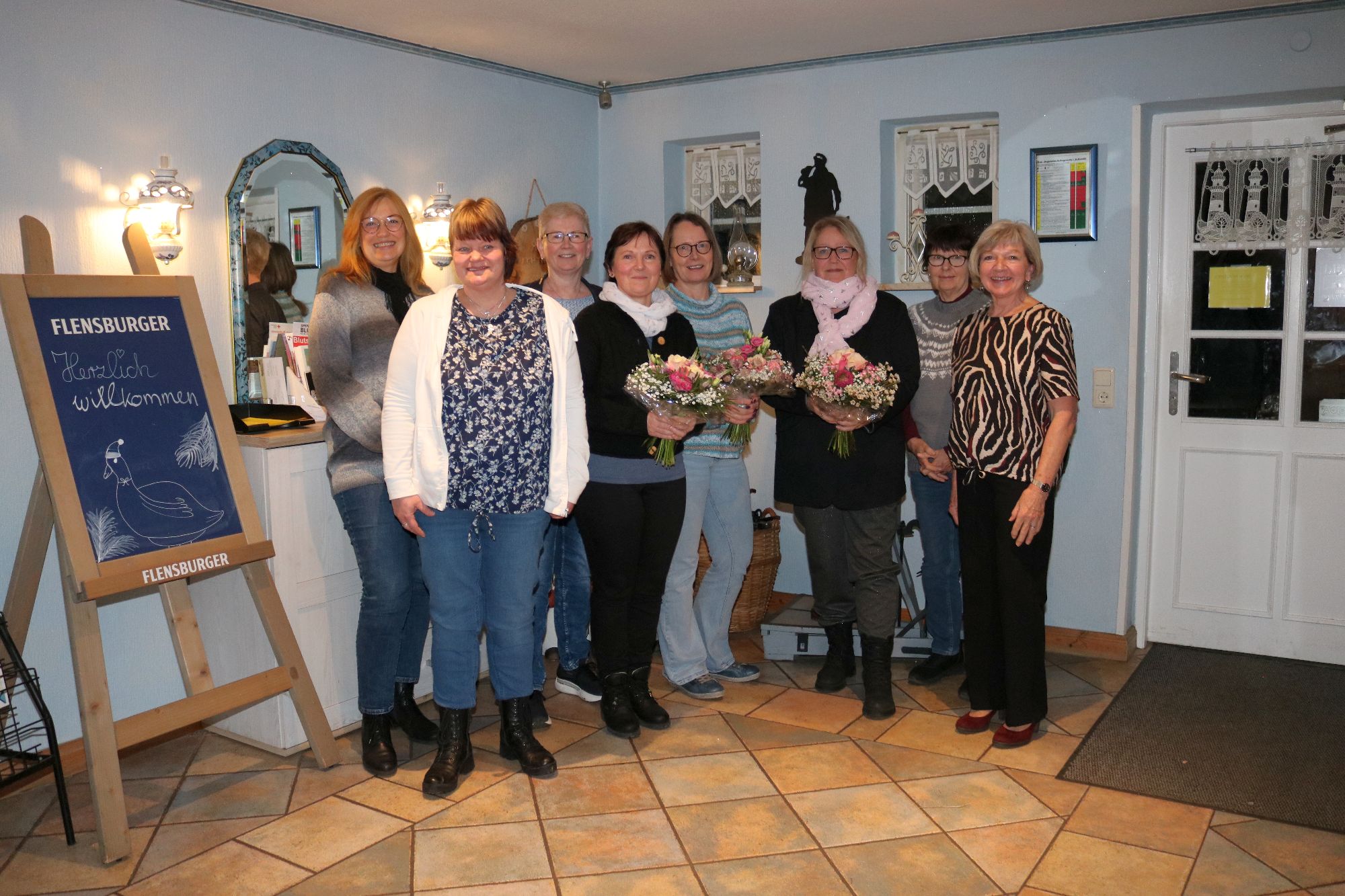 Acht Frauen stehen lächelnd im Eingangsbereich der Achtruper Stuben zusammen, zwei von ihnen halten Blumensträuße in der Hand. Links ein Kreidetafel-Aufsteller mit der Aufschrift „Herzlich willkommen".