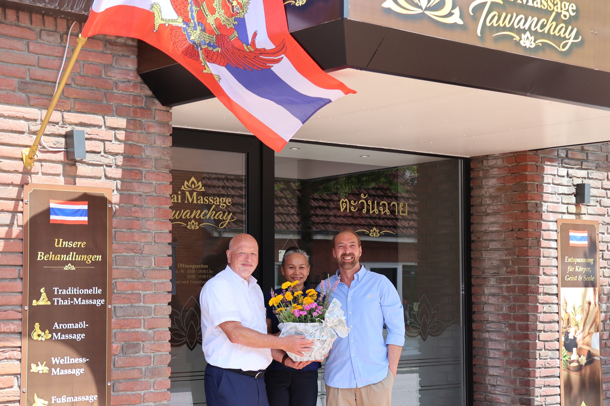 Bürgermeister Andreas Deidert überreicht Tawanchay Witt eine Blumenschale vor dem Eingang von Thaimassage Tawanchay in der Hauptstraße 31 in Leck, im Hintergrund eine thailändische Flagge.