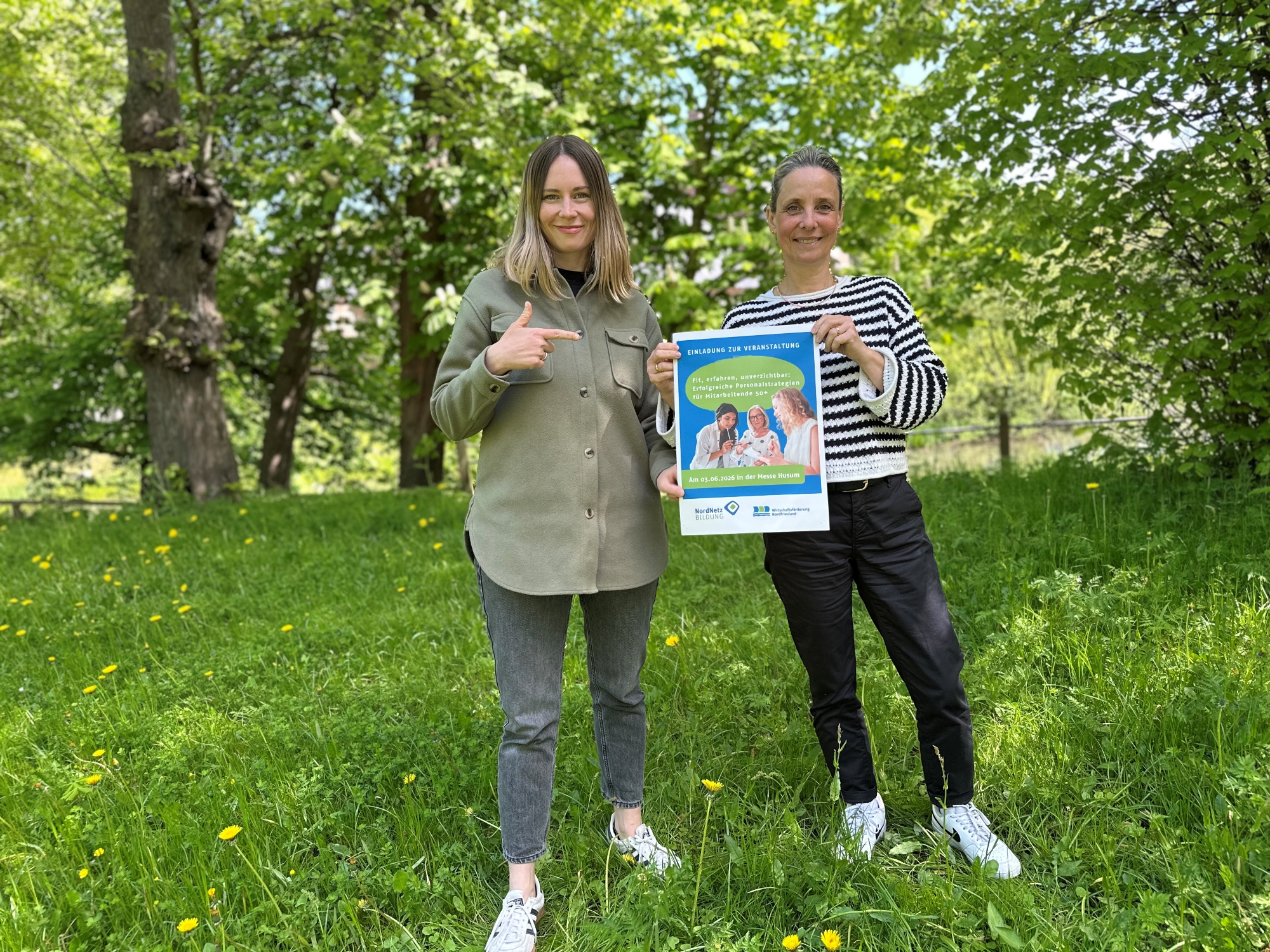 Stefanie Goelling und Christina König halten das Veranstaltungsplakat für „Fit, erfahren, unverzichtbar – Erfolgreiche Personalstrategien für Mitarbeitende 50+" in die Kamera.